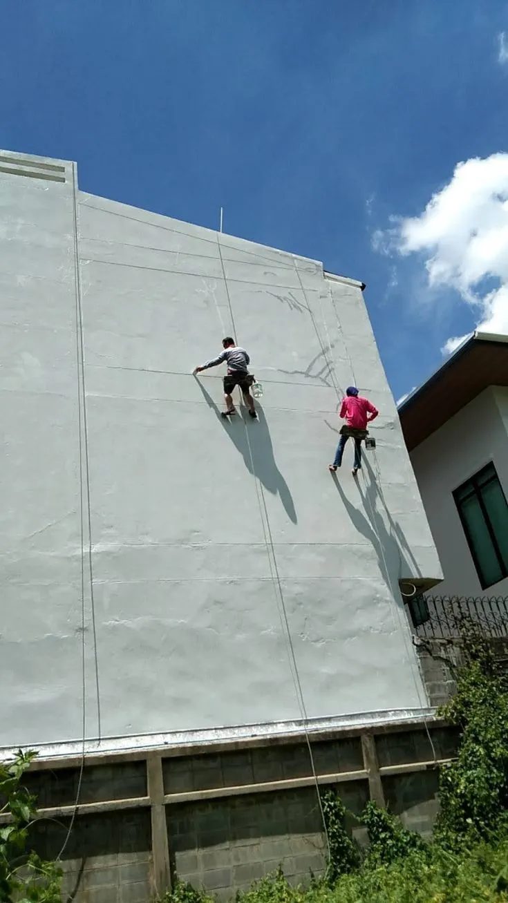 Two painters on ropes work down a tall, freshly coated exterior wall under a blue sky.